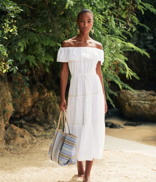 Woman in white dress on beach carries striped tote bag, accessorized with earrings. Verdant foliage and sand frame the scene.