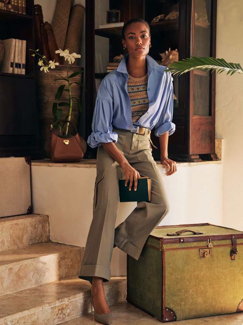 A woman poses indoors wearing an unbuttoned blue shirt, patterned top, khaki pants, and pumps. She holds a dark-green book and bag.