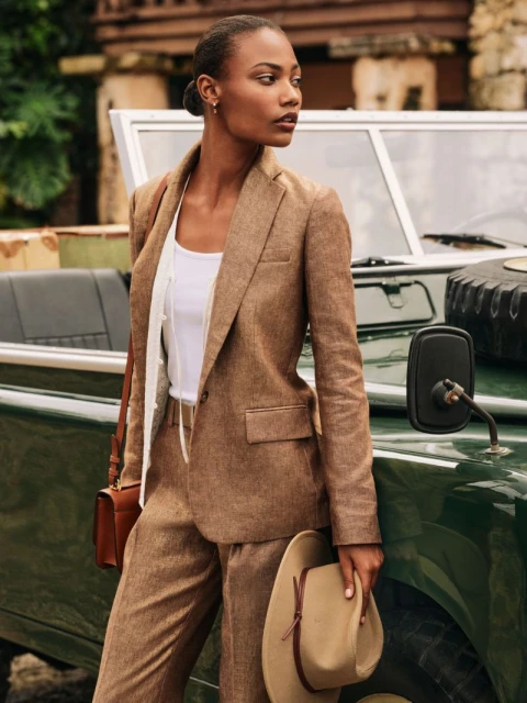 A stylish woman in a brown suit stands next to a vintage green SUV, holding a tan cowboy hat in an outdoor fashion setting.