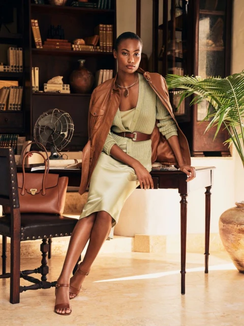 Elegant woman in light green dress with brown leather jacket, sitting on a desk with a leather handbag and vintage fan.