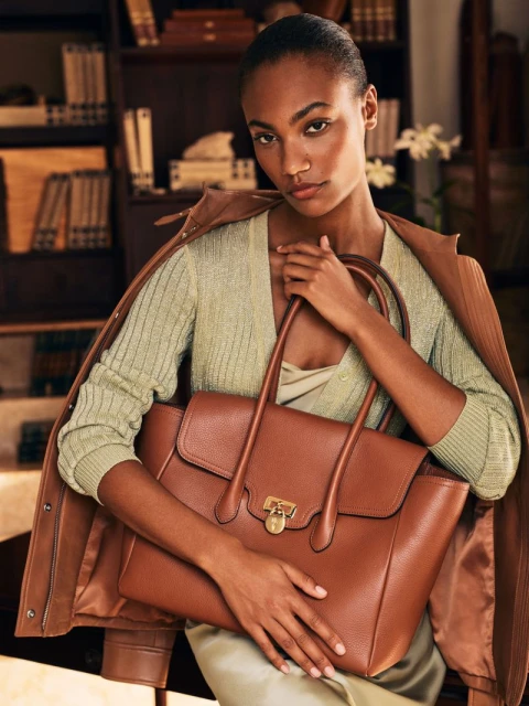 A woman with light brown skin poses indoors with a brown leather tote bag. She is wearing a light green sweater and brown jacket.