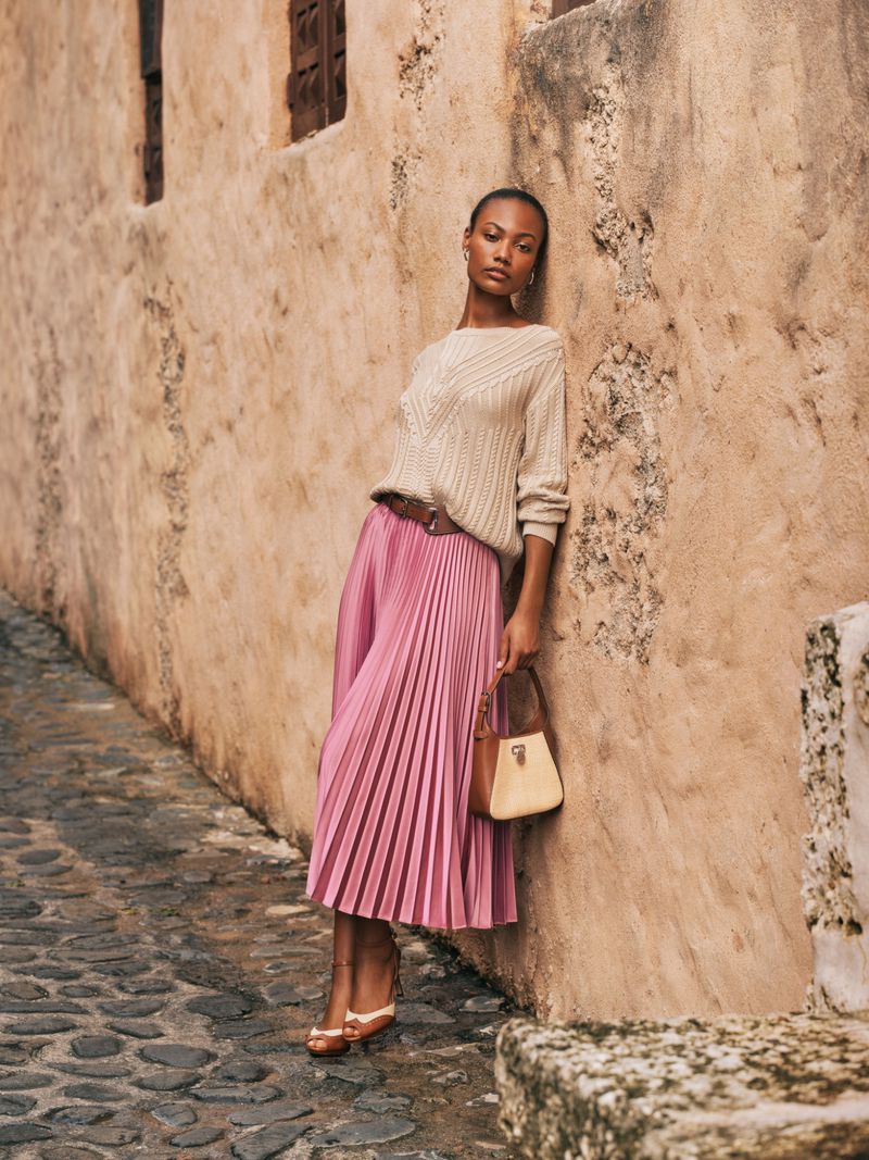 A woman in a sweater and pleated skirt leans against an old building on a cobblestone street with a small handbag in hand.