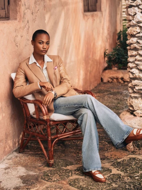 A poised Black woman in a tan blazer and jeans sits in a wicker chair outdoors against a stucco wall, creating a relaxed fashion portrait.