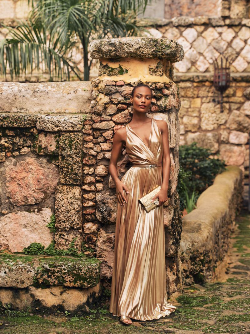 Elegant adult female in a gold gown poses near a stone wall outdoors, holding a matching clutch, fashion photoshoot.