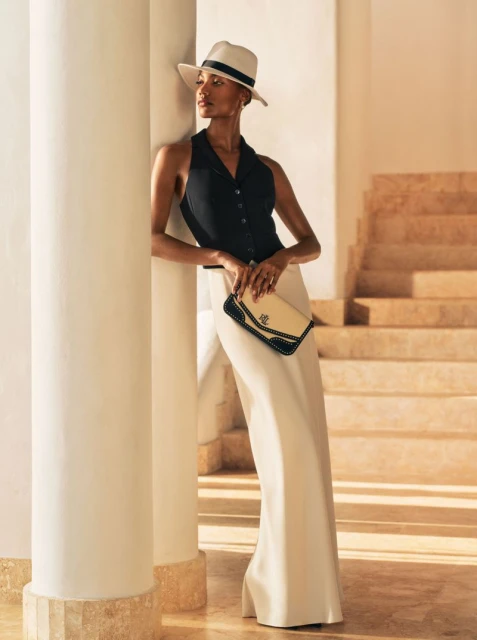 Elegant adult woman in black top and wide-legged pants poses with a clutch against columns and stairs in soft, diffused light.