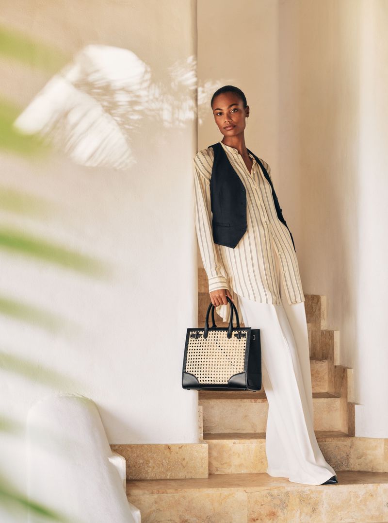 Fashionable Black woman in a striped shirt and vest poses with a handbag on stairs. Warm lighting, neutral colors, and indoor setting.