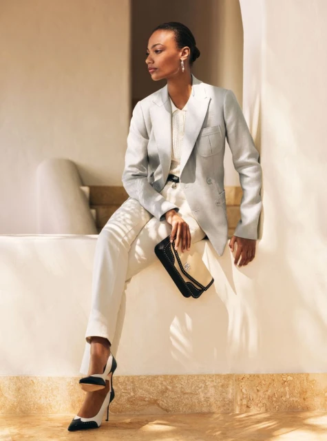 Elegant woman in light-grey blazer and white pants sitting, holding a clutch. Black and white heels, white wall background.
