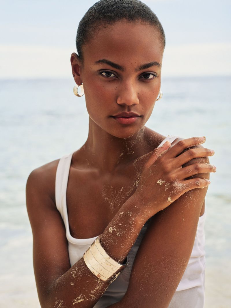Portrait of an adult woman with tan skin and short hair, wearing hoop earrings and a bracelet, posed on a sandy beach with an ocean backdrop.
