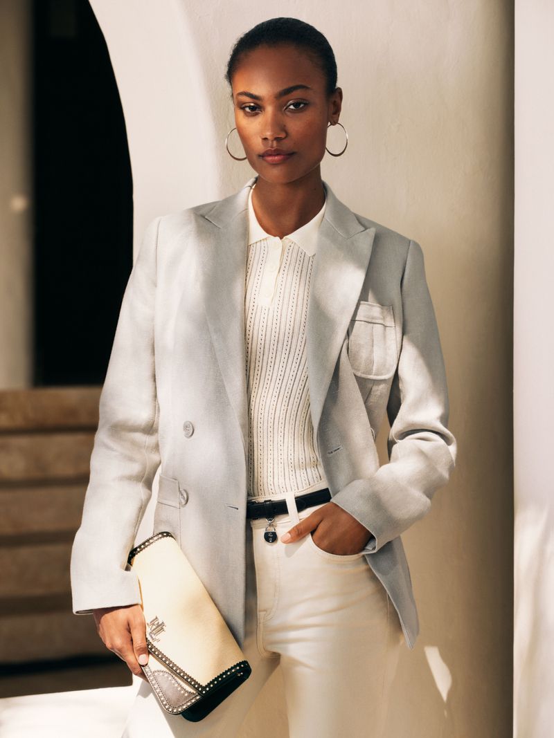 Elegant woman in a grey blazer, cream top and pants poses indoors, holding a clutch. The fashion portrait features soft, diffused lighting.