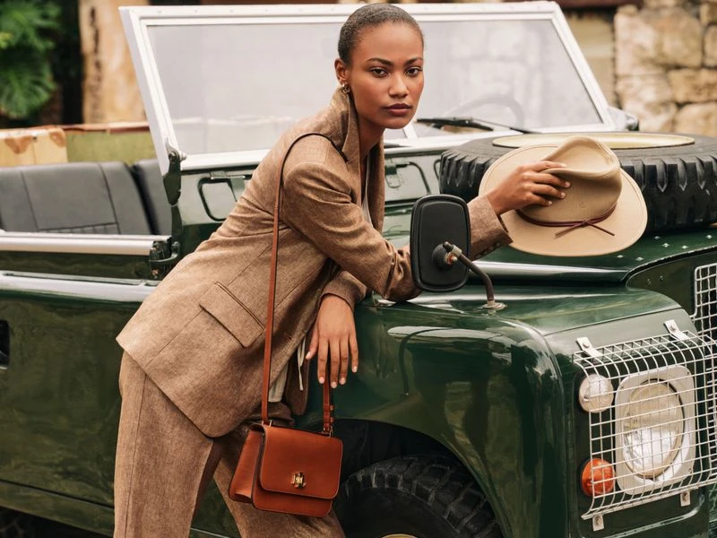 Woman in tweed suit poses by a vintage Land Rover; she holds a hat in hand, with a tan leather satchel hanging at her side.