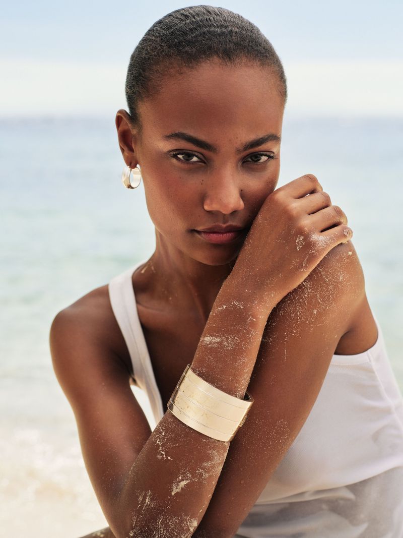 Model with short dark hair wearing a white tank top and gold cuff bracelet against a soft ocean background