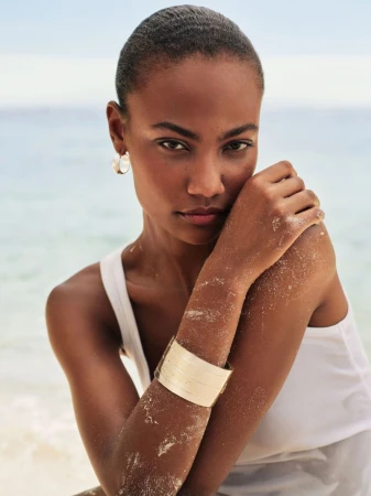 Model with short dark hair wearing a white tank top and gold cuff bracelet against a soft ocean background