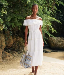 Model in white off-the-shoulder dress holding a striped tote bag on a sandy beach