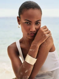Model with short dark hair wearing a white tank top and gold cuff bracelet against a soft ocean background