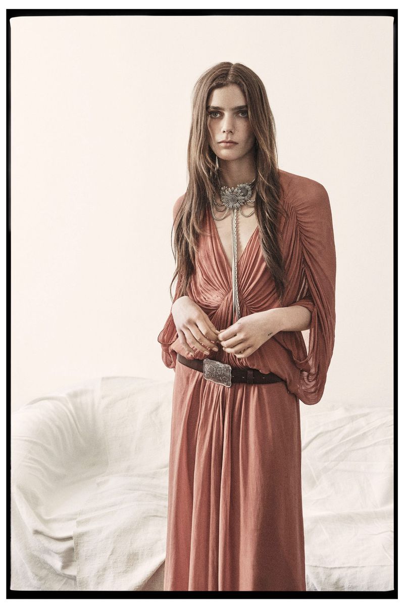 An adult female fashion model poses in a flowing peach-colored dress and belt with a white sofa in the background against a neutral wall.