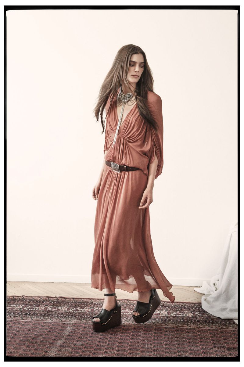 A fashionable woman with long brown hair wearing a brown maxi dress, black belt, and black platform shoes stands on an oriental rug.