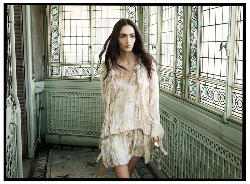 A woman with long dark hair stands in a light-filled hallway wearing a floral dress and gold necklace, looking to the side.