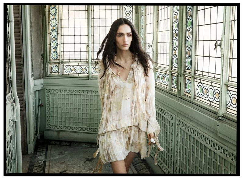 A woman with long dark hair stands in a light-filled hallway wearing a floral dress and gold necklace, looking to the side.