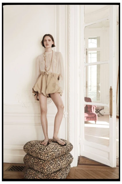 A young woman poses on a stack of patterned cushions in a fashion shot, wearing beige balloon shorts and a sheer blouse in a white room.