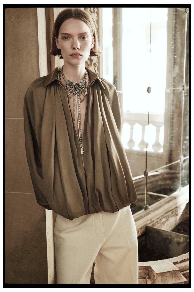 Adult female fashion model posing indoors wearing oversized button-down shirt, pants, and ornate necklace in a natural light fashion editorial.