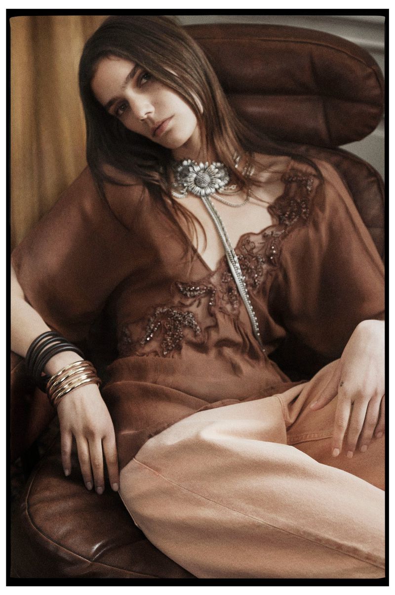 Brunette woman in brown blouse and tan pants sits in leather chair wearing bolo tie and bracelets in a portrait-style fashion shot.