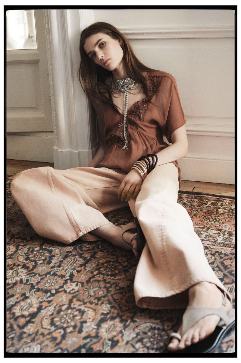 A female model poses indoors wearing a brown blouse, wide peach pants, and jewelry, sitting on a patterned rug in a neutral-toned room.