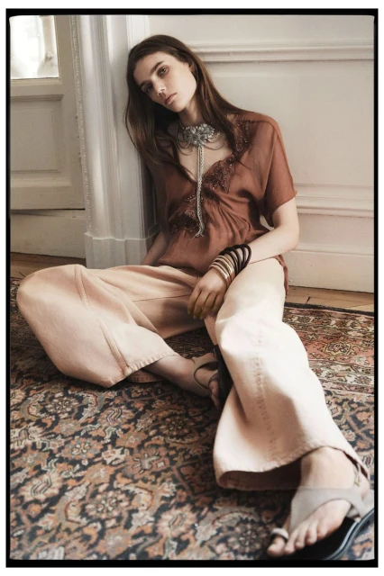 A female model poses indoors wearing a brown blouse, wide peach pants, and jewelry, sitting on a patterned rug in a neutral-toned room.