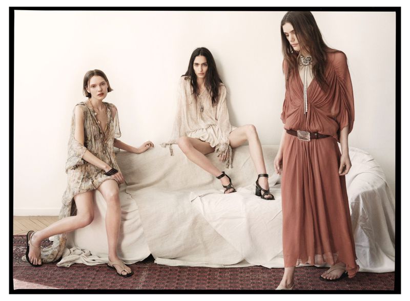 Three women pose in dresses on a couch in a fashion editorial. The woman in the middle sits with legs spread apart.