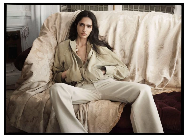 A brunette woman wearing a beige jacket and pants sits on a couch with a floral-patterned blanket in an indoor setting.