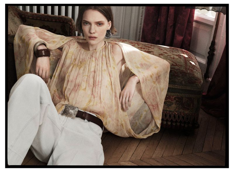 A woman in a flowing floral blouse and white jeans is posed in a relaxed position in a room with a vintage-style daybed.