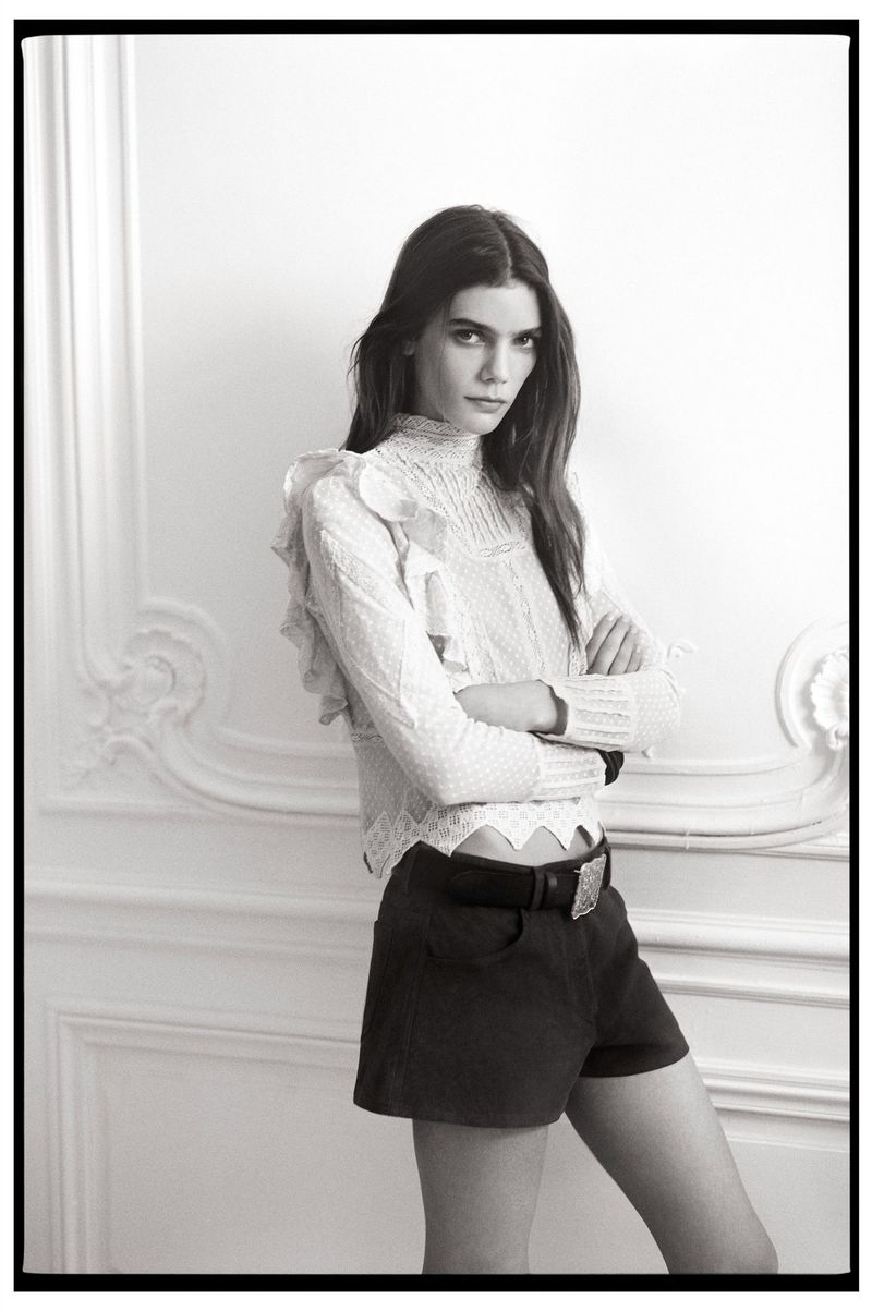 A woman poses in a white ruffled top and dark shorts against a white wall with architectural details in a monochrome fashion shot.