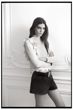 A woman poses in a white ruffled top and dark shorts against a white wall with architectural details in a monochrome fashion shot.