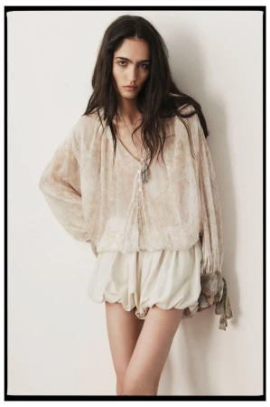 Indoor shot of a young adult woman in a printed blouse and shorts, posing against a neutral wall with soft, diffused lighting.