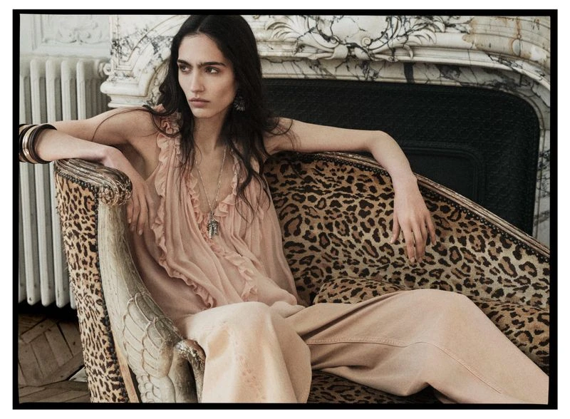 A woman in a pink dress sits on a leopard print chaise lounge with a marble fireplace in the background in an indoor setting.