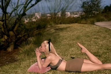 A woman lying on a grassy hill outdoors, propped on a pink pillow with blonde hair and a black bow.