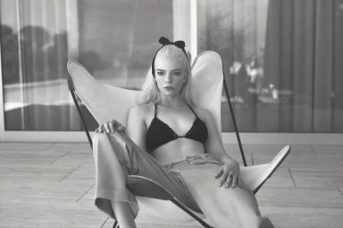 Anya Taylor-Joy seated in a butterfly chair wearing a dark halter top and bow headband against a modern background.
