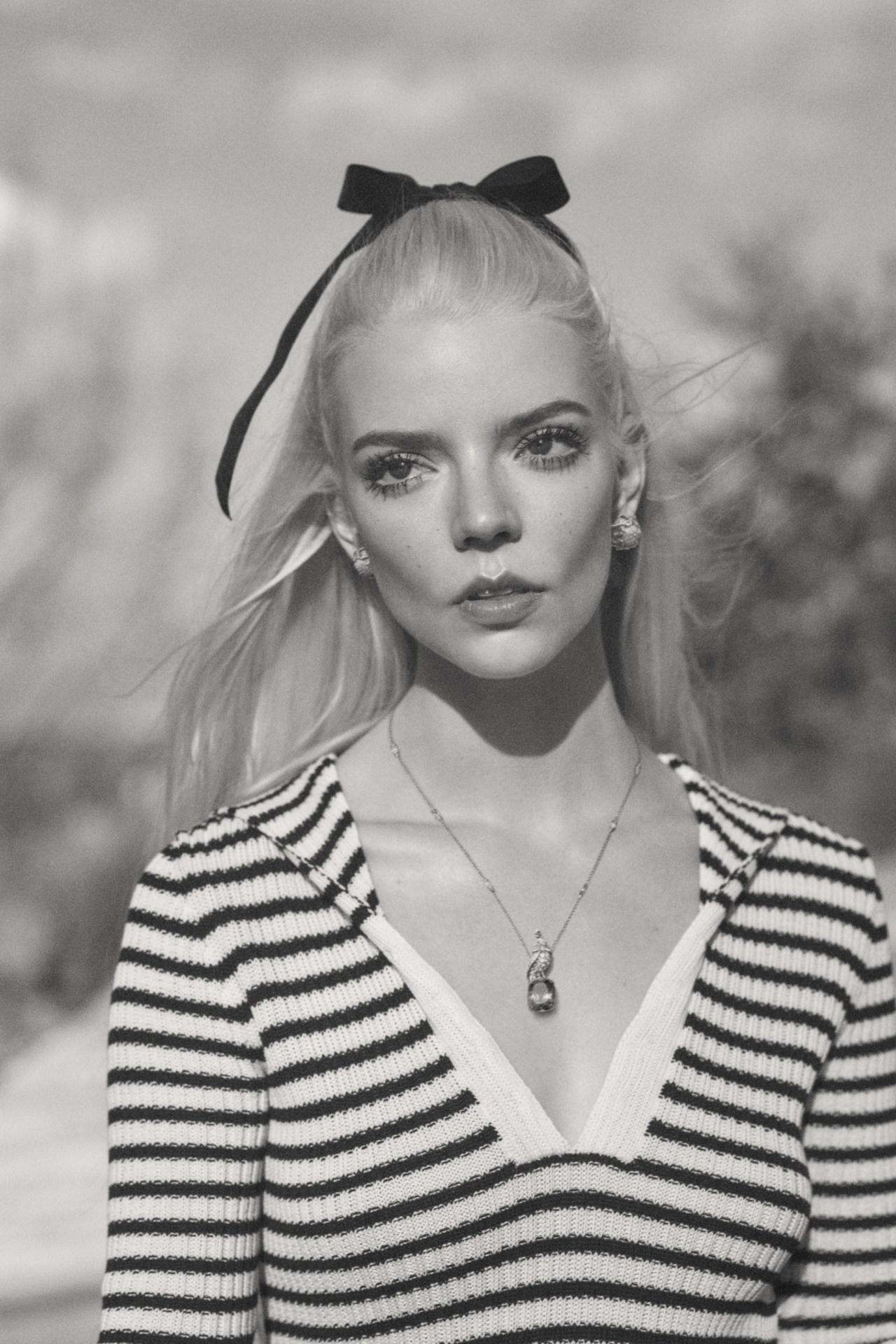 Black-and-white portrait of Anya Taylor-Joy with a black bow in her platinum blonde hair, wearing a striped shirt outdoors.
