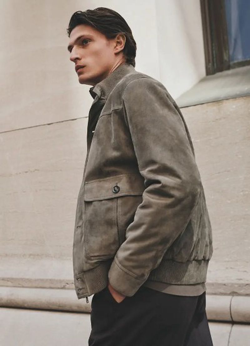 Man in stylish suede bomber jacket stands near a building, captured in a medium shot with soft, natural lighting, showcasing fashion.