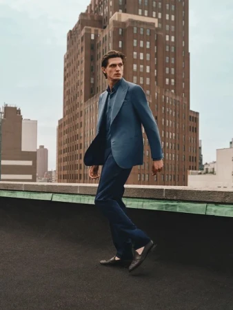 Man in blue suit walks on rooftop with city buildings in background on overcast day, fashion editorial style, outdoor setting.