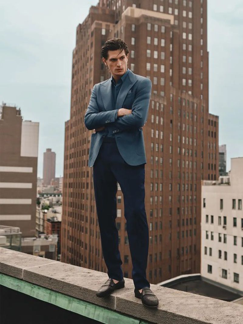 Man standing with arms crossed wearing a blue blazer and dark trousers on a rooftop with skyscrapers behind him.