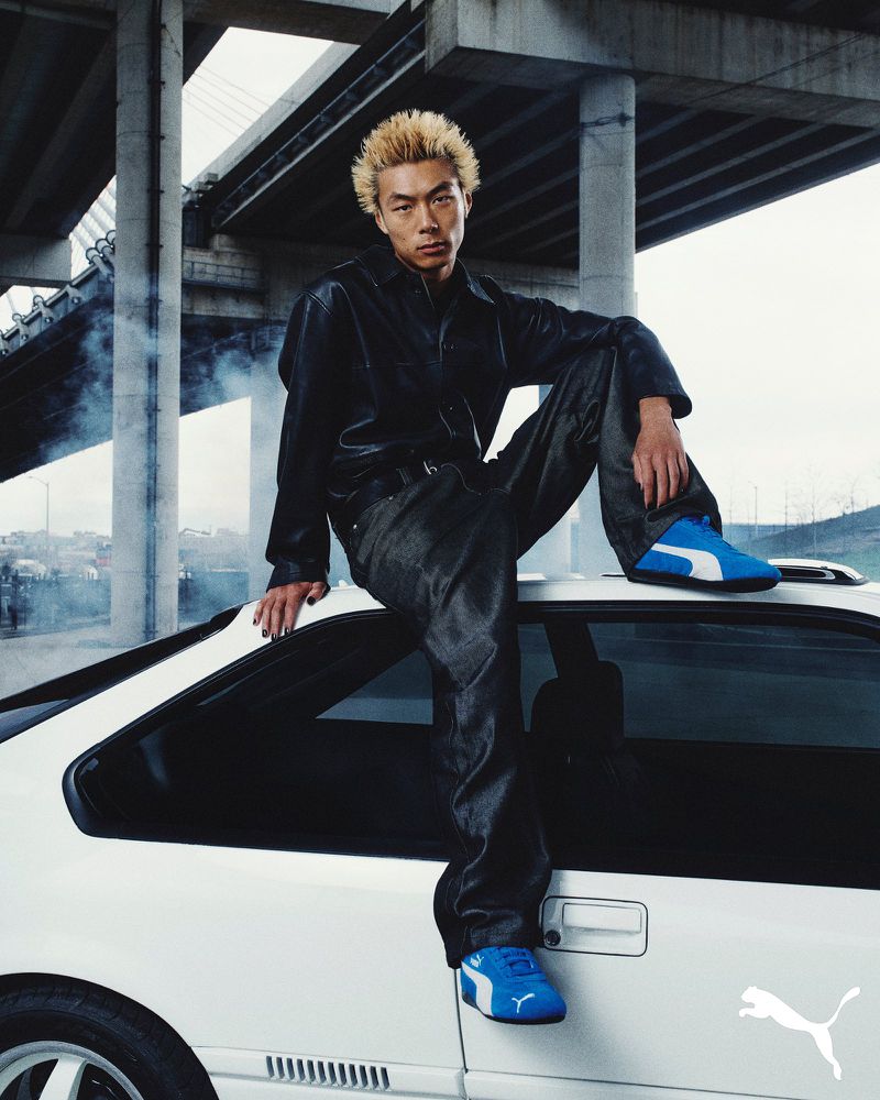 Asian man in dark denim jacket sits on white car with blue Puma sneakers; urban overpass structure in background with vapor effect.