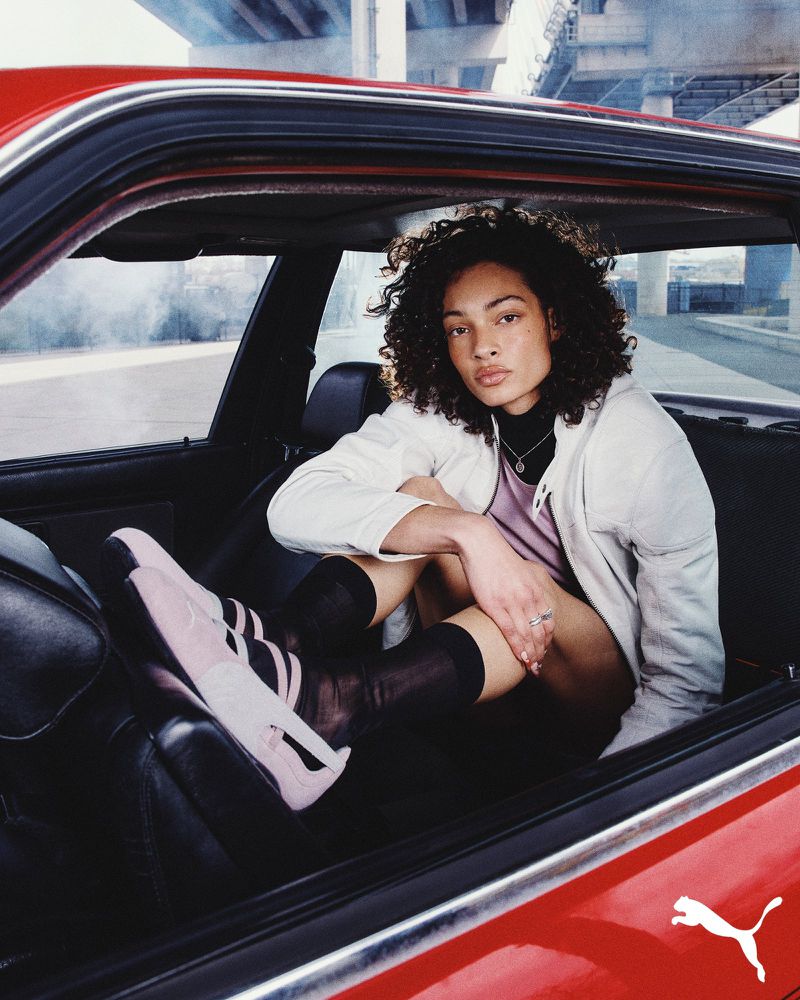 An adult female poses inside a red car, wearing pink Puma sneakers, knee-high socks, and a white jacket, with an urban background.