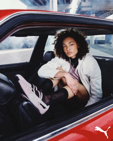 An adult female poses inside a red car, wearing pink Puma sneakers, knee-high socks, and a white jacket, with an urban background.