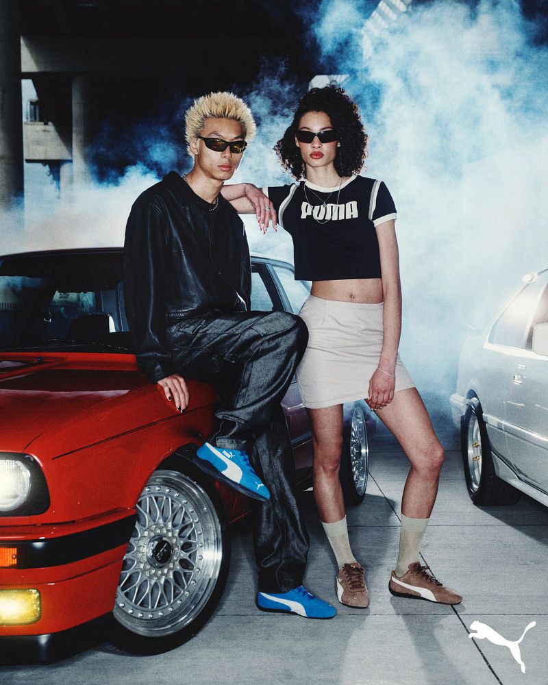 Two young adults pose near classic BMW cars in a stylized fashion shoot, showcasing streetwear and PUMA shoes in an urban environment.