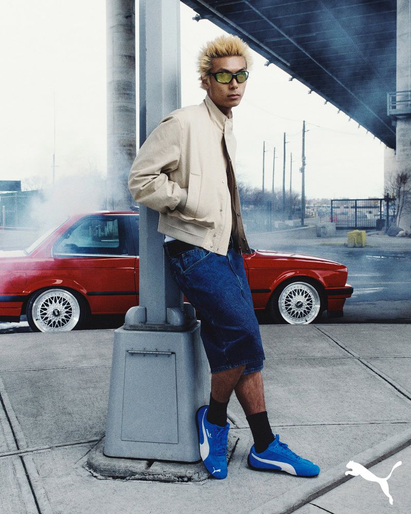 Man in a beige jacket leans against a gray pillar near a red car under a bridge. He wears blue shoes and has blond hair.