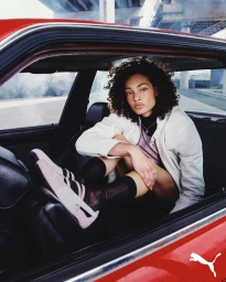Model with curly hair sitting inside a red car wearing sneakers, styled by Marcus Correa for Puma The Speedcat.