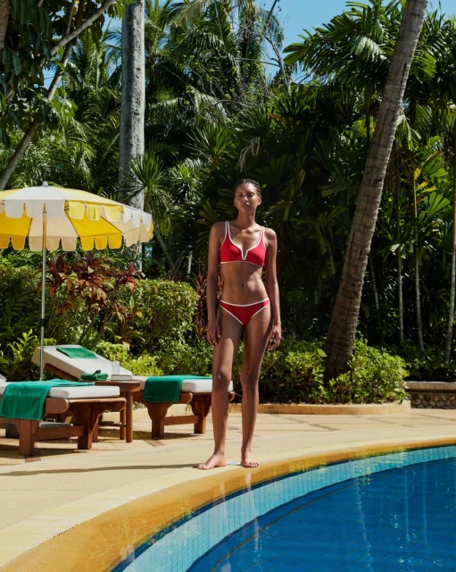 Woman in a red bikini poses by a blue pool with green tropical foliage and palm trees in the background on a sunny day outdoors.