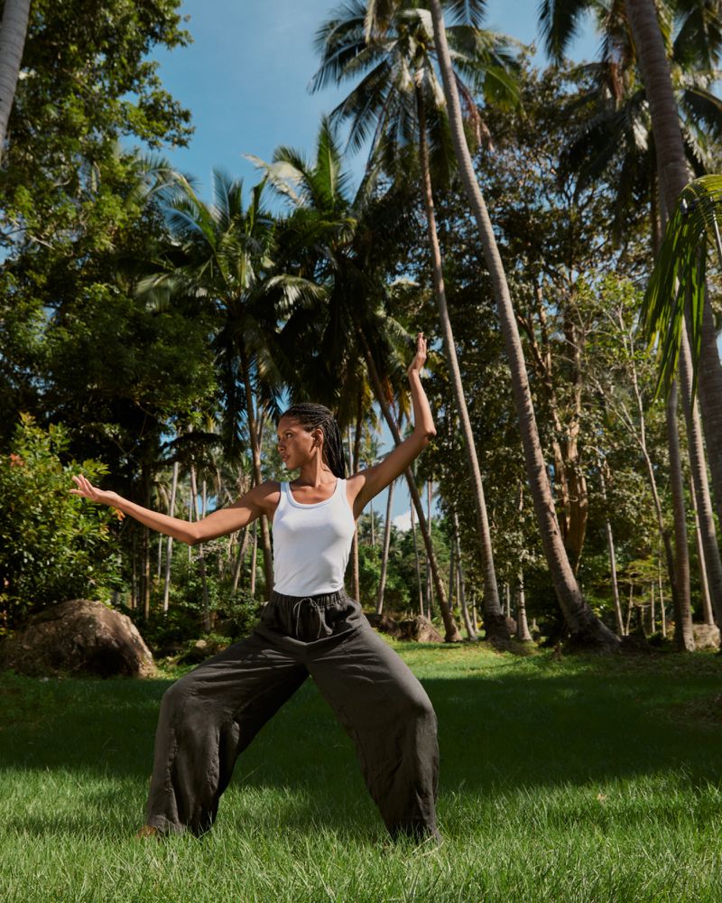Adult woman in white tank top and wide leg pants balances on a grass lawn with palm trees and forest background during daytime.