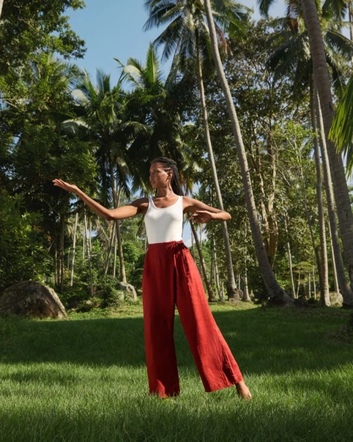 Woman in red pants stands in lush green field with palm trees, striking artistic pose with arms outstretched in natural sunlight.