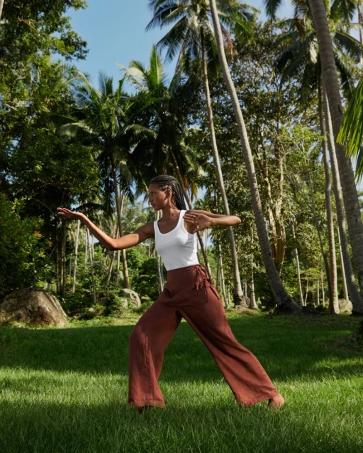 Woman in loose brown pants and a white top performs a yoga pose on a lawn surrounded by palm trees and lush green foliage.